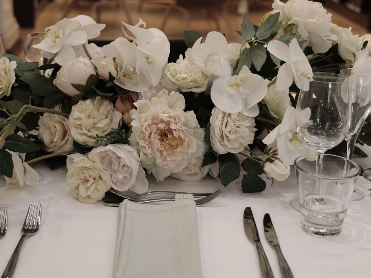 Table de mariage dressée, chemin de fleurs blanches et de verdure, verres et couverts alignés sur la nappe