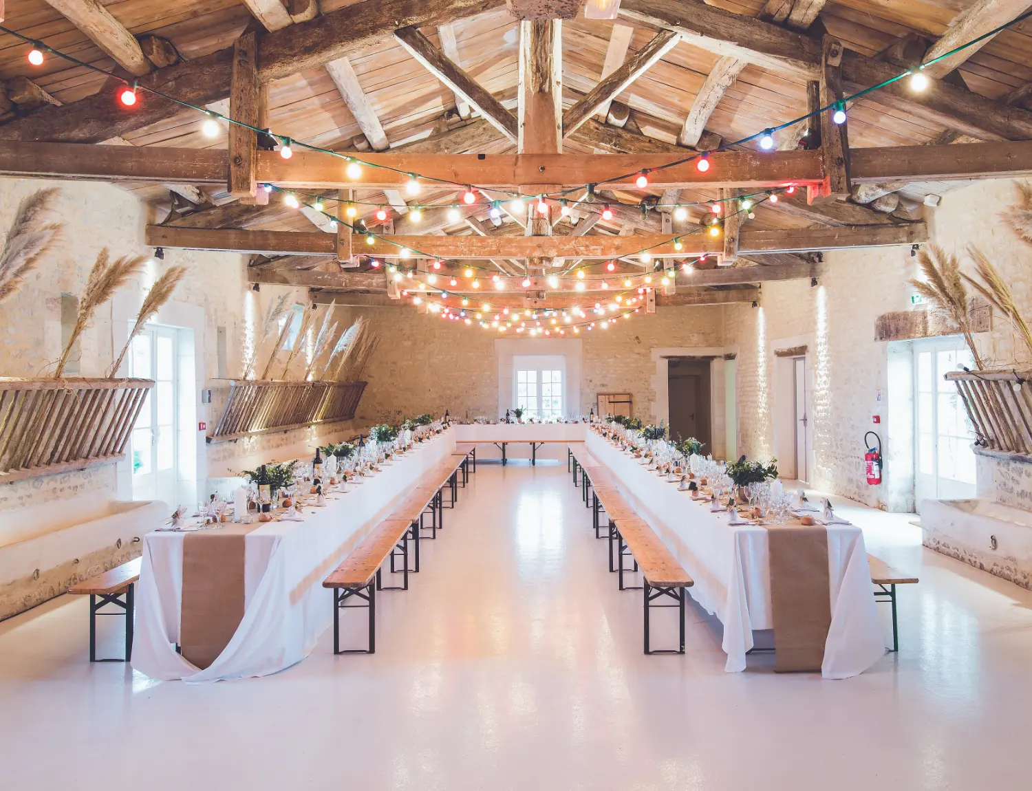 Grande salle de réception dressée pour un banquet, longues tables nappées de blanc et guirlandes lumineuses au plafond