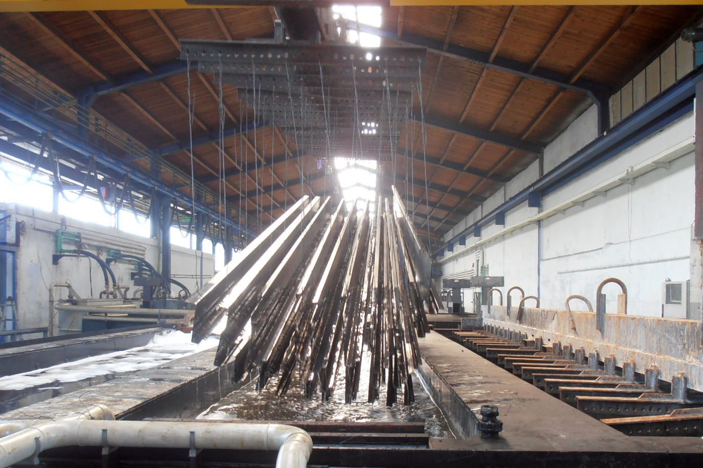 Poutres d’acier suspendues à un palonnier, sortant du bain de zinc en fusion d’une galvanisation à chaud, dans une halle couverte.