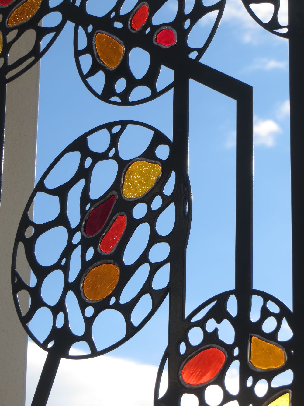 Panneau d’acier découpé au laser en motifs ajourés, garni de verres colorés jaune, orange et rouge, photographié en contre-jour sur ciel bleu.
