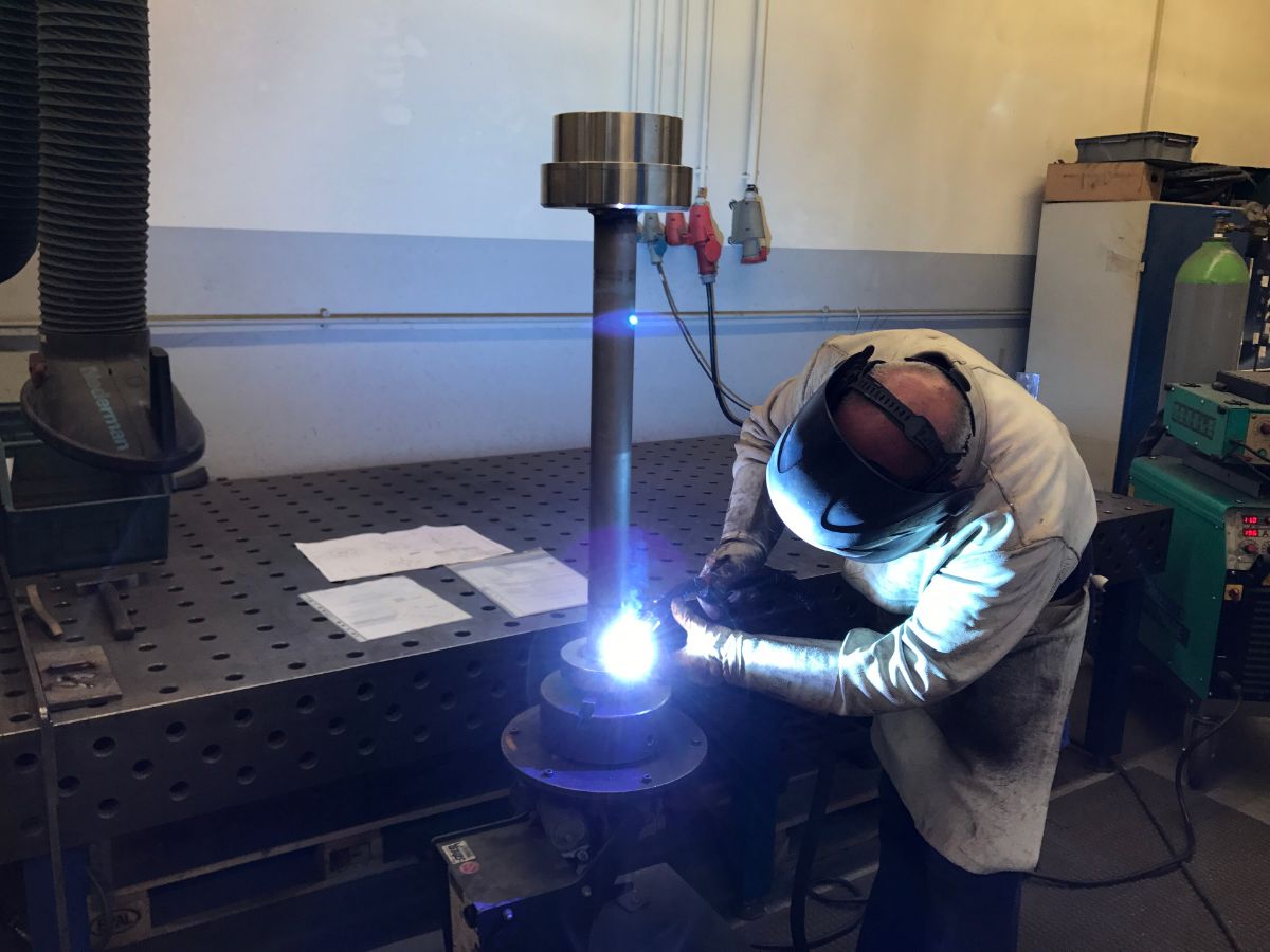 Soudeur casqué en train de souder une bride sur une pièce tubulaire, posée sur une table de soudage perforée, arc bleu et bras aspirant au-dessus.
