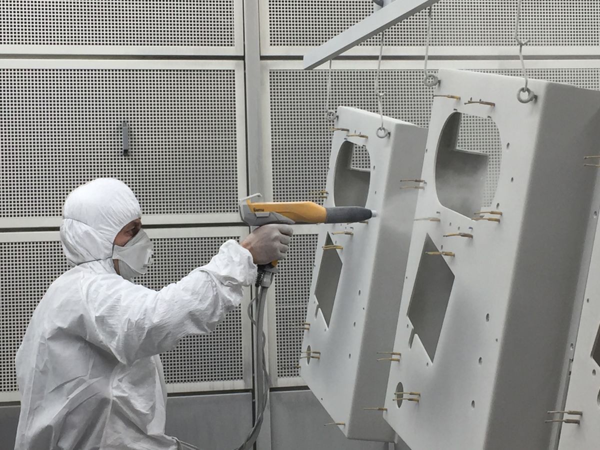 Opérateur en combinaison blanche appliquant de la poudre au pistolet électrostatique sur des panneaux de tôle suspendus dans une cabine de thermolaquage.