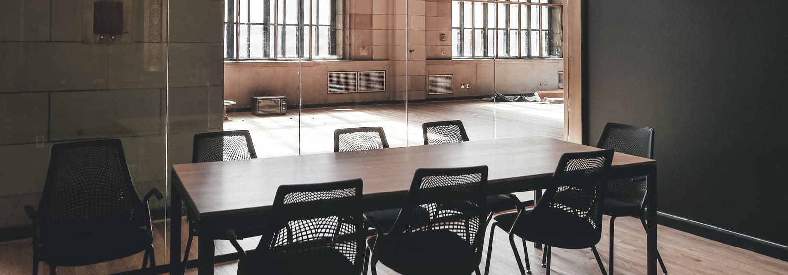 Une salle de travail vide, longue table en bois, chaises noires et grandes fenêtres d'atelier.