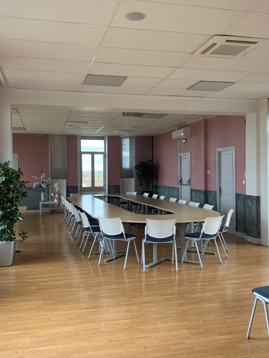 Salle du conseil municipal vide, tables disposées en fer à cheval, chaises pour le public alignées au fond