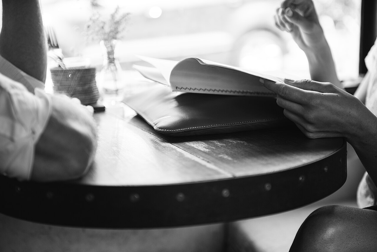 Deux personnes assises de part et d'autre d'une petite table, l'une tend un dossier à l'autre, photographie en noir et blanc