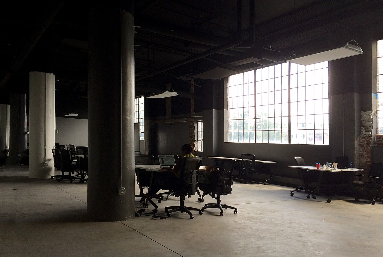 Un plateau de bureaux aux grandes fenêtres, plusieurs postes de travail inoccupés, photographie en noir et blanc