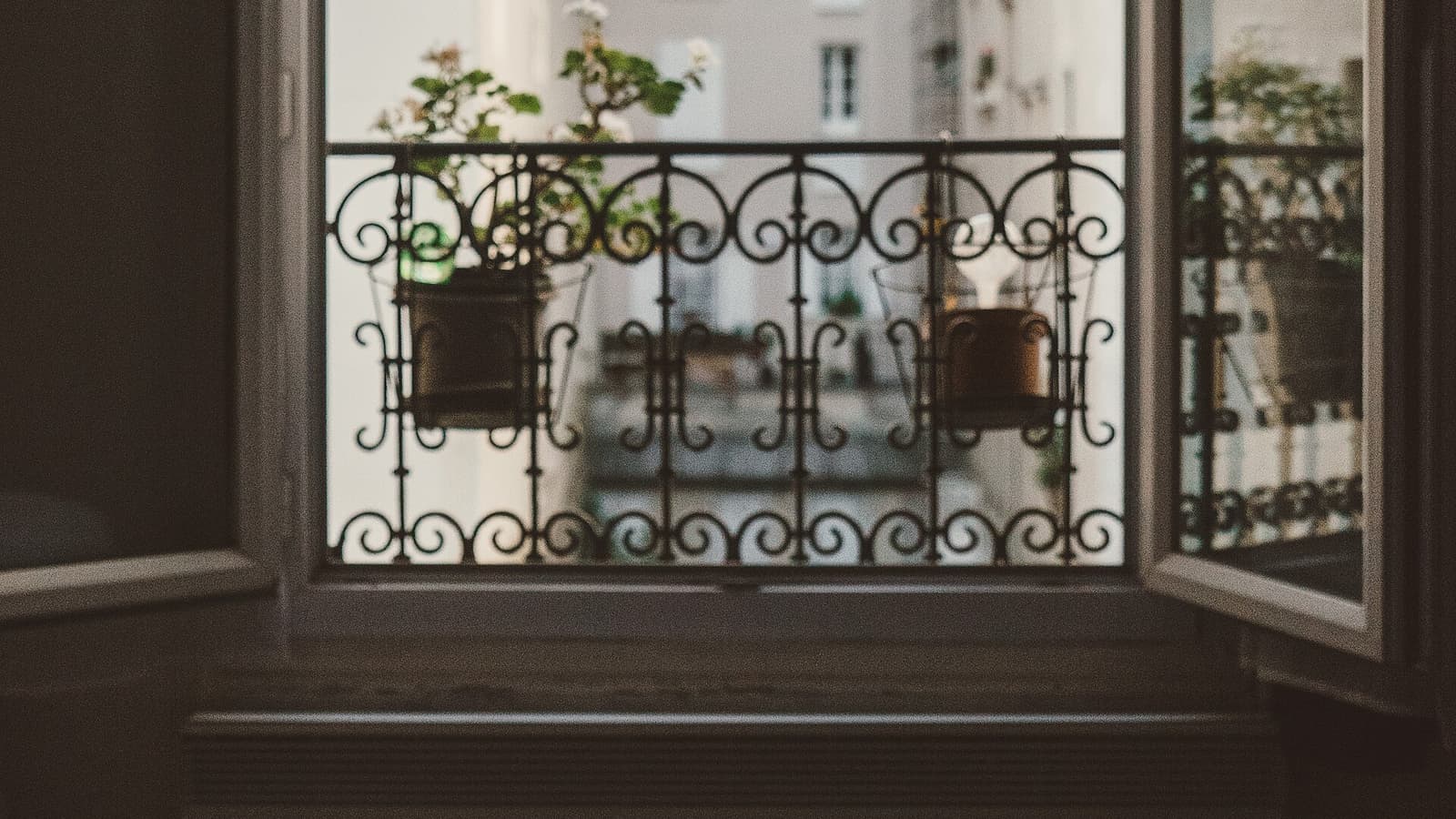 Fenêtre à deux battants ouverte sur un balcon en fer forgé fleuri, vue sur la façade claire d'en face, lit au premier plan