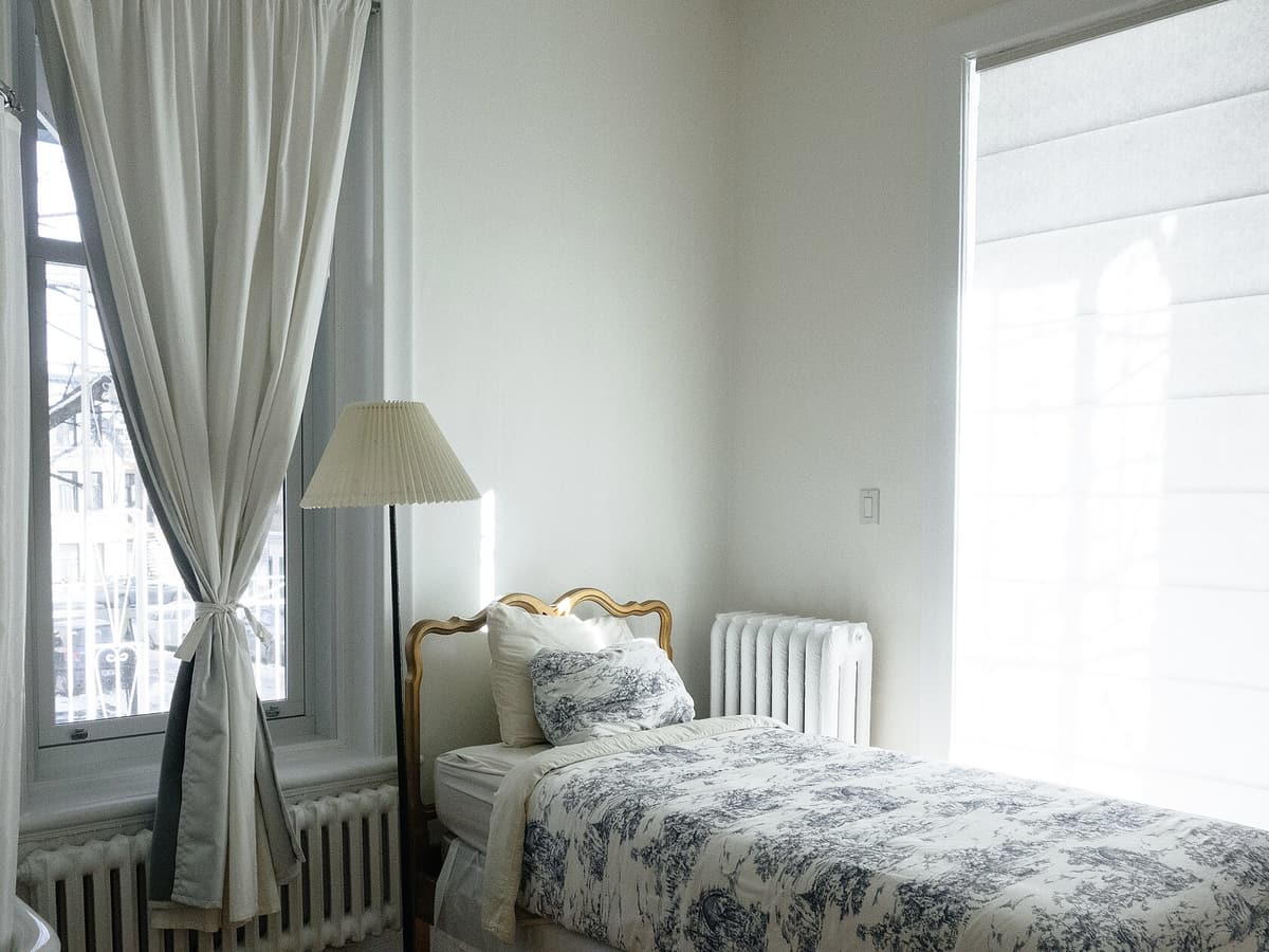 Chambre claire, lit simple à tête de lit dorée, couvre-lit à motif bleu, rideau de lin et radiateur en fonte devant la fenêtre