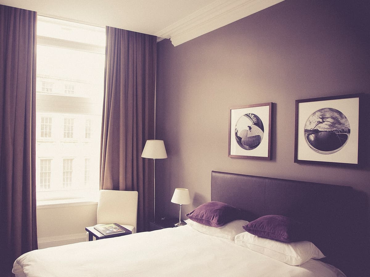 Chambre double, grand lit blanc, murs aubergine, hautes fenêtres à rideaux longs donnant sur une façade en pierre