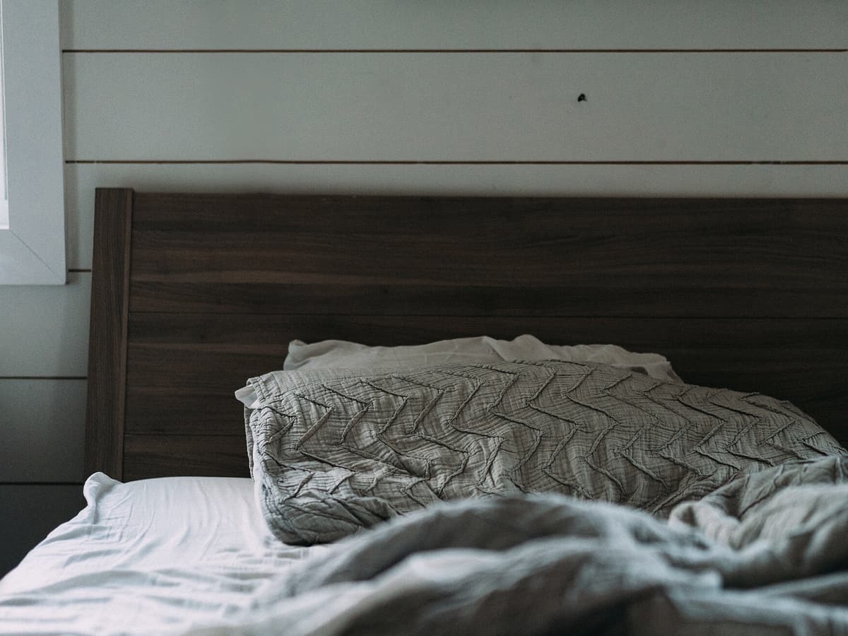 Lit large à tête de lit en noyer, plaid gris et draps blancs, mur lambrissé clair et fenêtre basse sur le côté