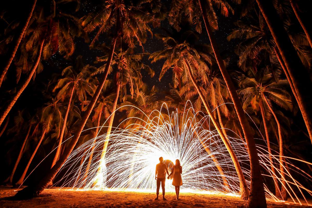 Deux silhouettes de loin sous des palmiers, gerbe d'étincelles dorées derrière elles dans la nuit