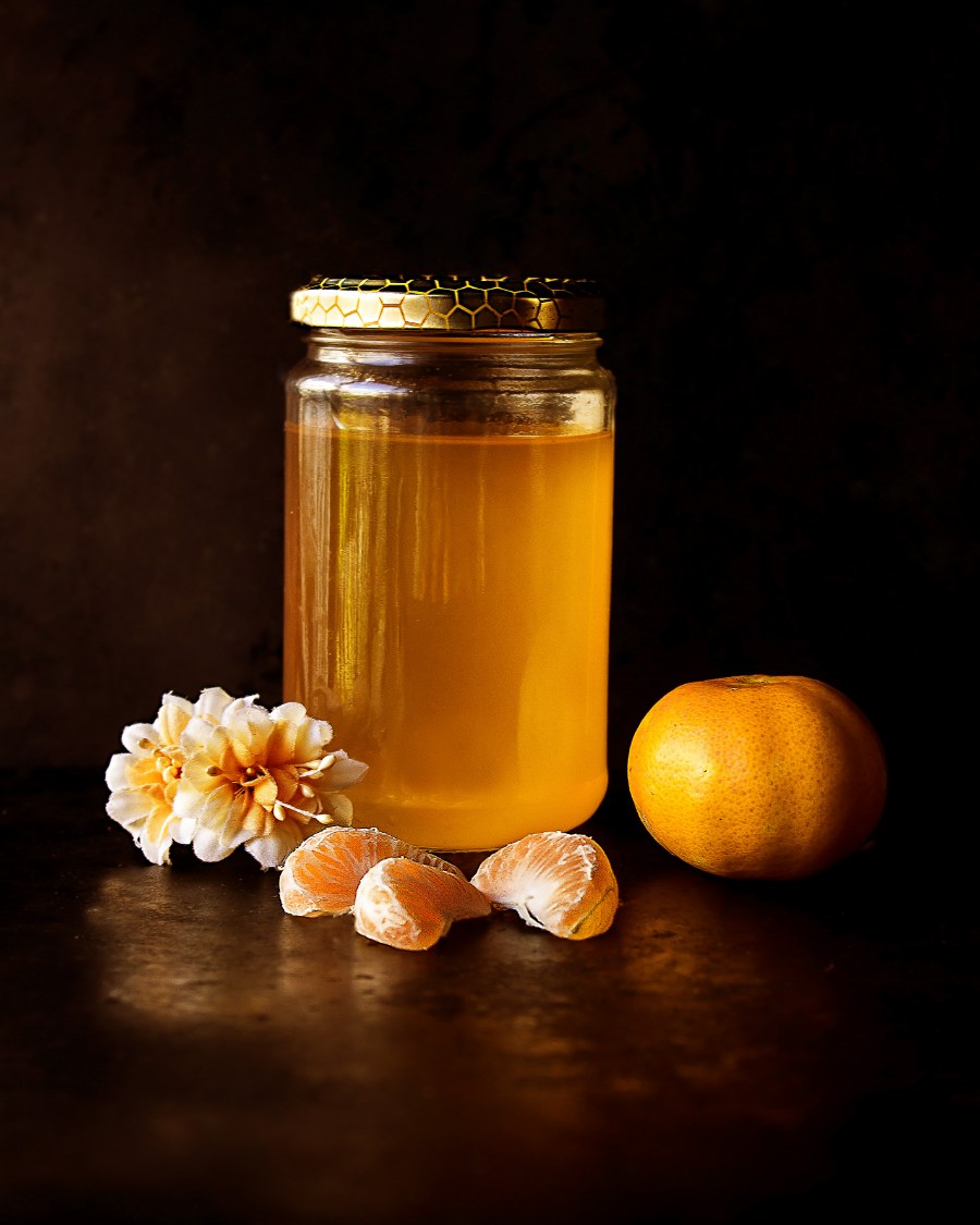Bocal de miel éclairé en lumière rasante sur fond noir, clémentine et fleurs séchées à côté