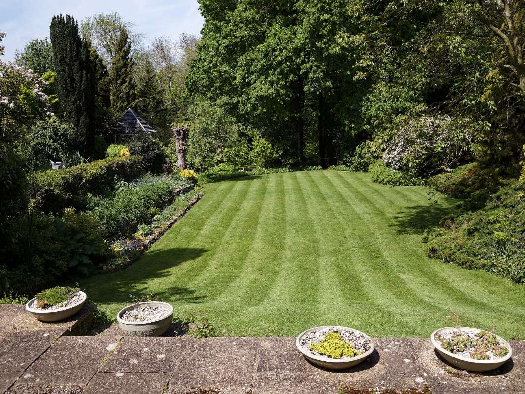 Jardin de particulier vu depuis la terrasse, grande pelouse tondue en bandes, massifs plantés en bordure et haies taillées.