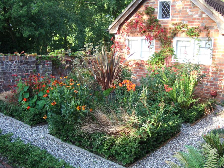 Massif planté de graminées et de vivaces orange, paillé de gravier gris, au pied d'une maison en brique.
