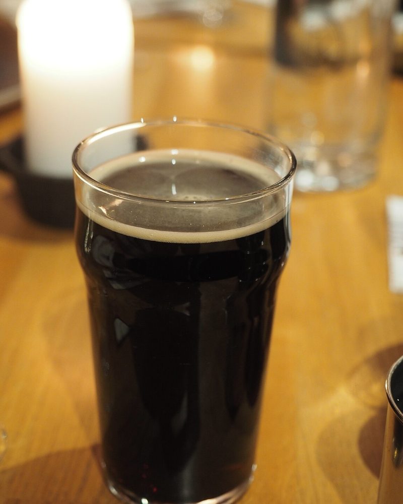 Verre de bière brune très foncée, mousse beige, sur une table en bois