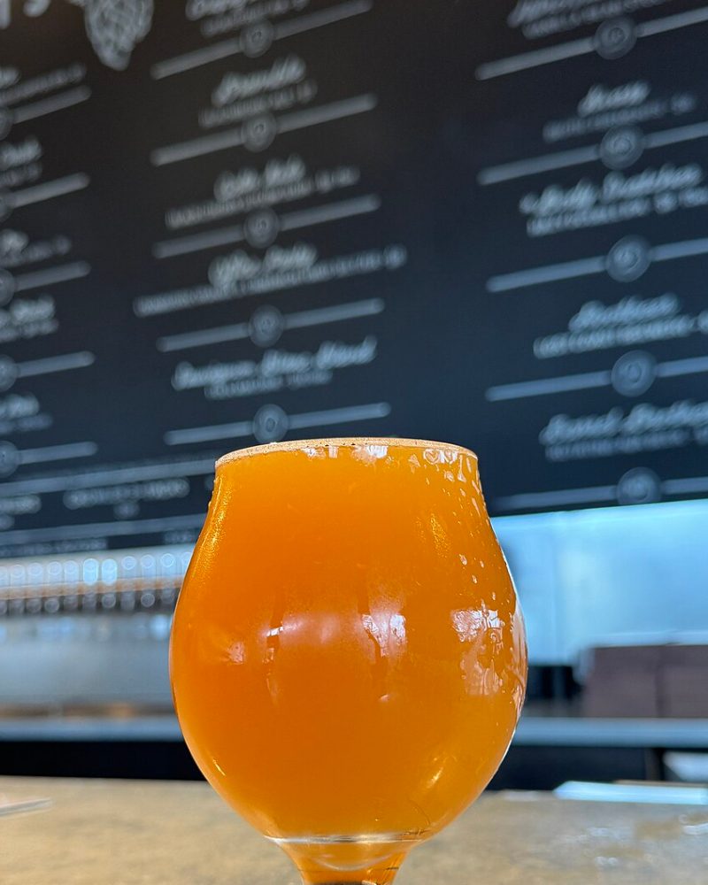 Verre tulipe rempli d'une bière orangée trouble, devant l'ardoise de la brasserie